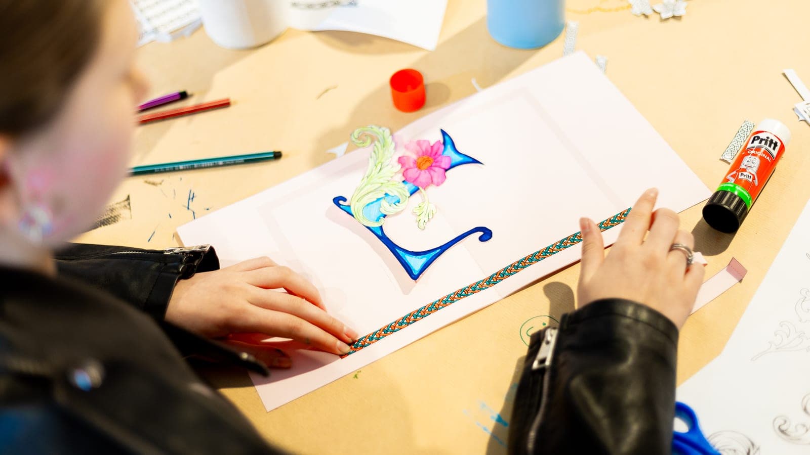 Pop-up cards workshop at Kulturhuef Musée — a child decorating a card with a hand-drawn initial, flower and decorative ribbon, with felt-tip pens and a glue stick laid out on the worktable.