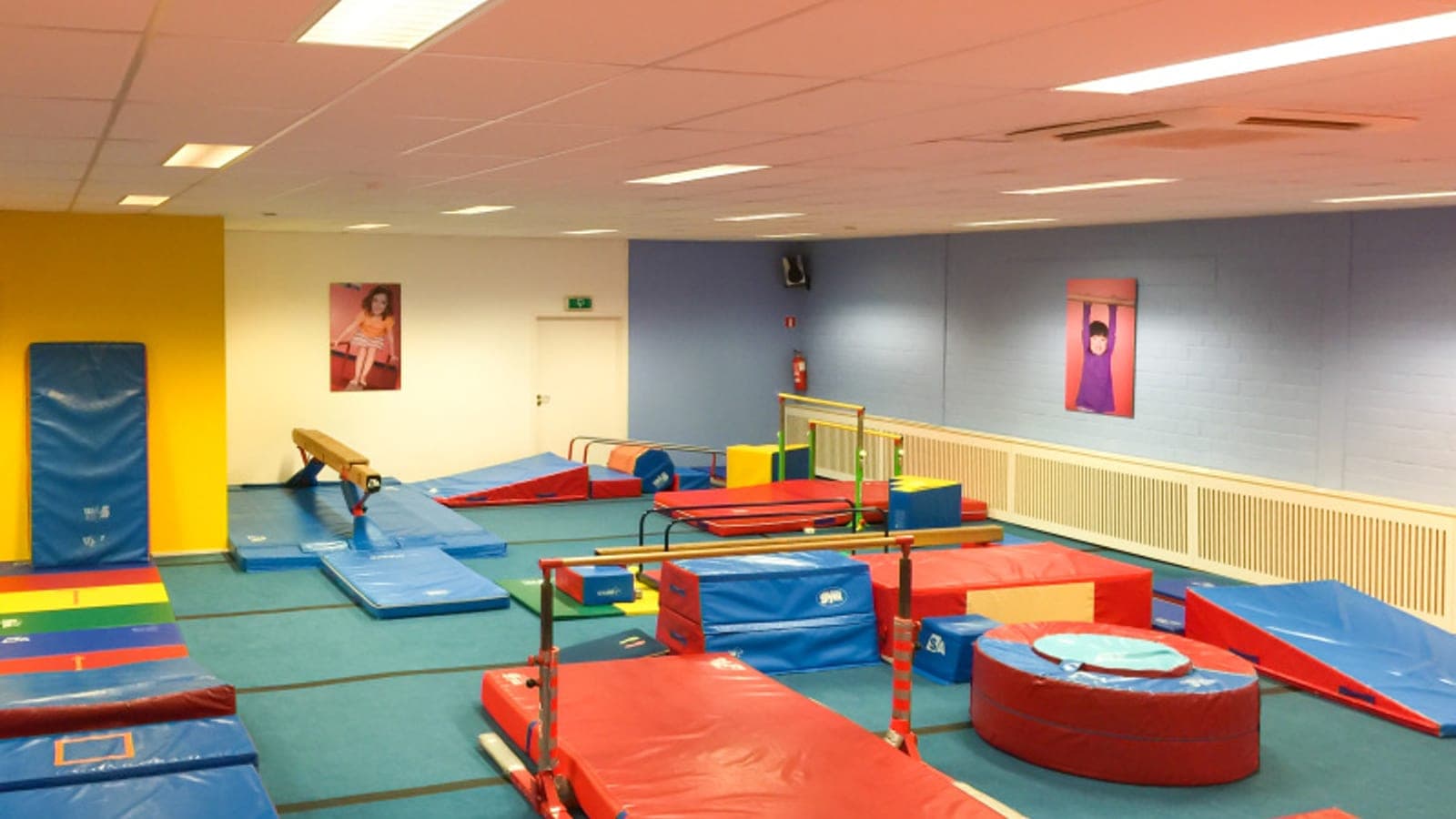 Wide view of The Little Gym Luxembourg gymnastics floor — vault, balance beam, parallel bars and coloured landing mats laid out across the room