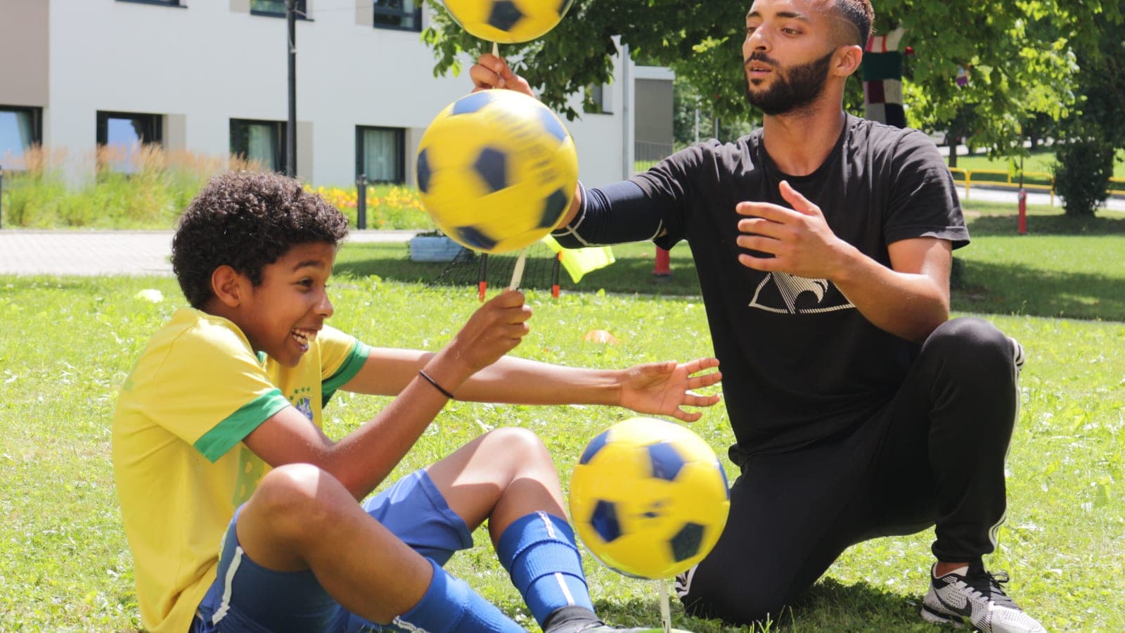 Mouloud crouched outdoors balancing three footballs around a child wearing a Brazil jersey.